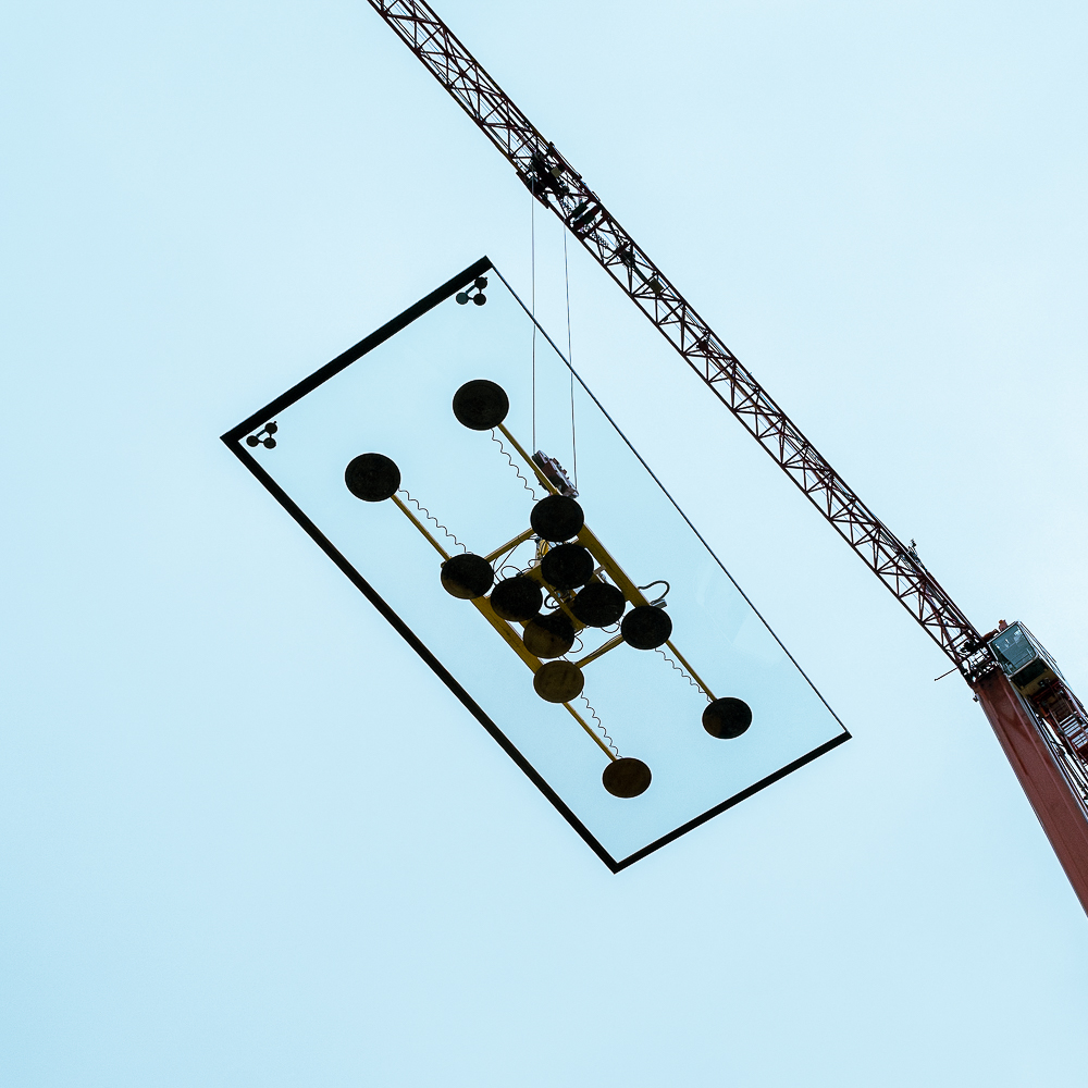A crane lifts a large glass panel using suction cups against a clear sky.