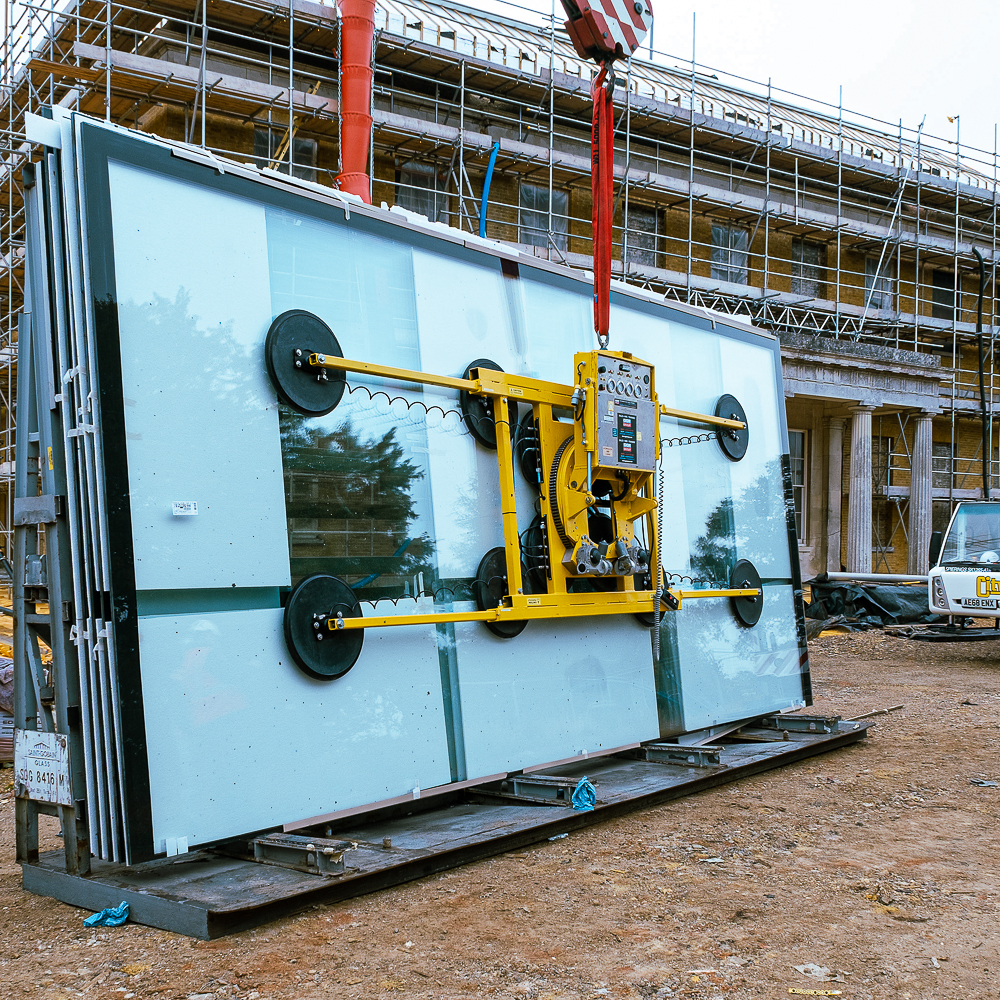 A vacuum lifter holds large glass panels at a construction site, with scaffolding and a building in the background.