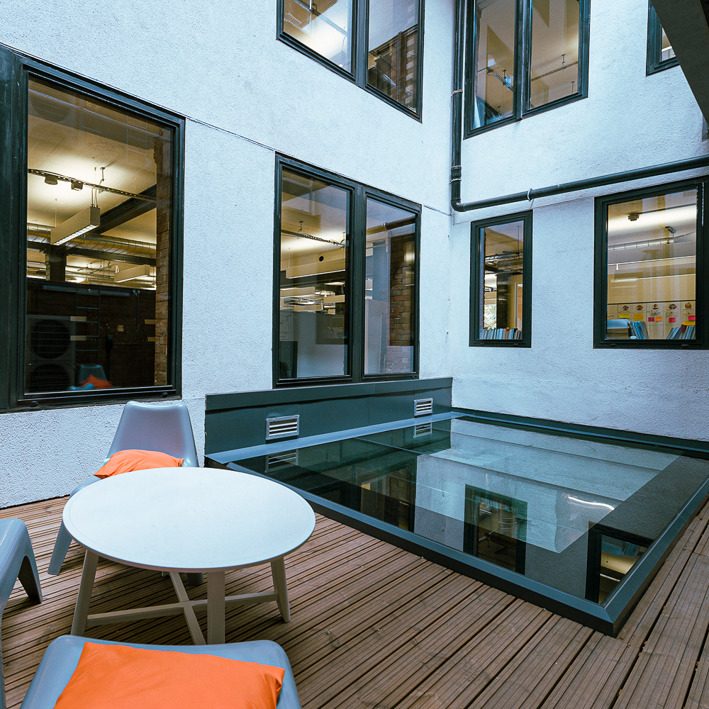 Modern outdoor space with a square glass-bottom feature surrounded by wooden decking, white table, gray chairs, and orange cushions, adjacent to a building with multiple windows.
