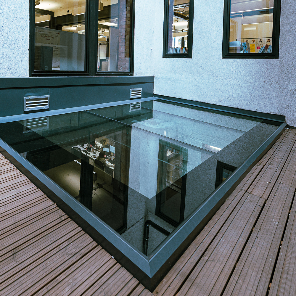 Glass floor with reflections, surrounded by wooden decking, outside modern building windows.