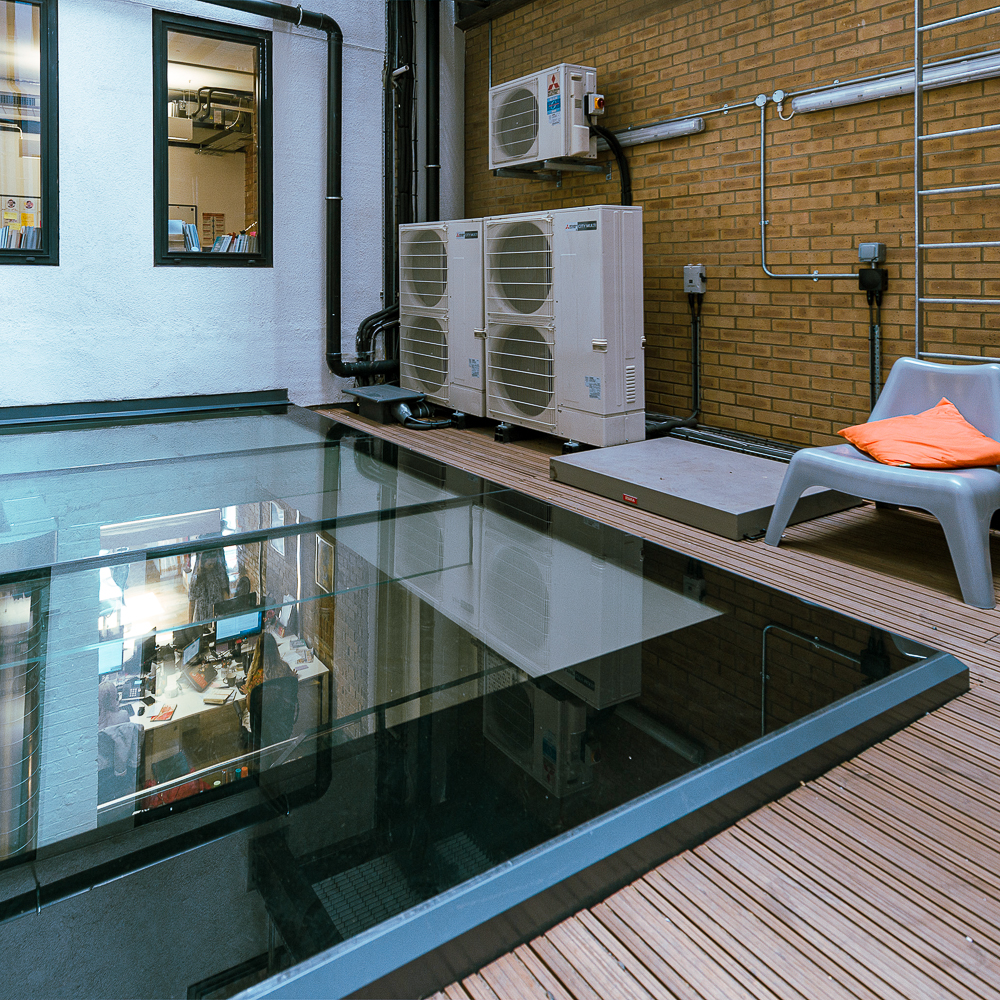 A modern room with a glass floor revealing an office below. Outdoor seating with a gray chair and orange cushion. Air conditioning units and brick walls are visible.
