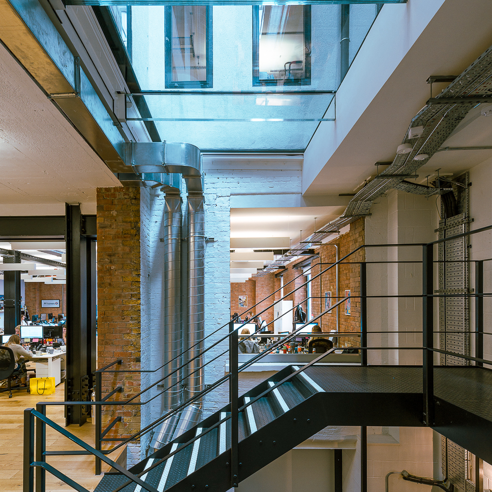 Modern industrial office interior with exposed brick walls, metal staircase, and visible ductwork. Bright lighting from overhead fixtures and windows enhances the open space.