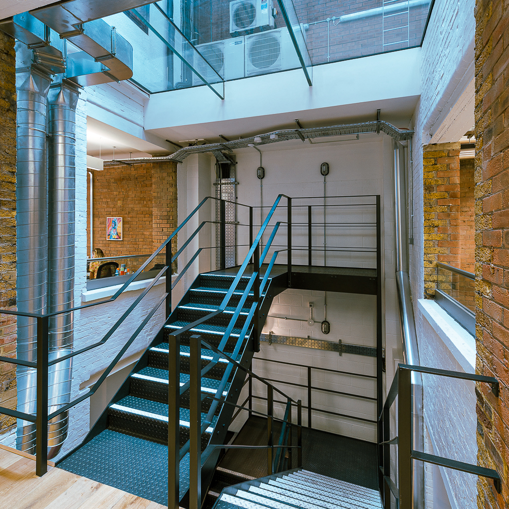Industrial-style staircase with black railings and perforated steps in a modern building. Brick walls and large glass windows are visible in the surroundings.