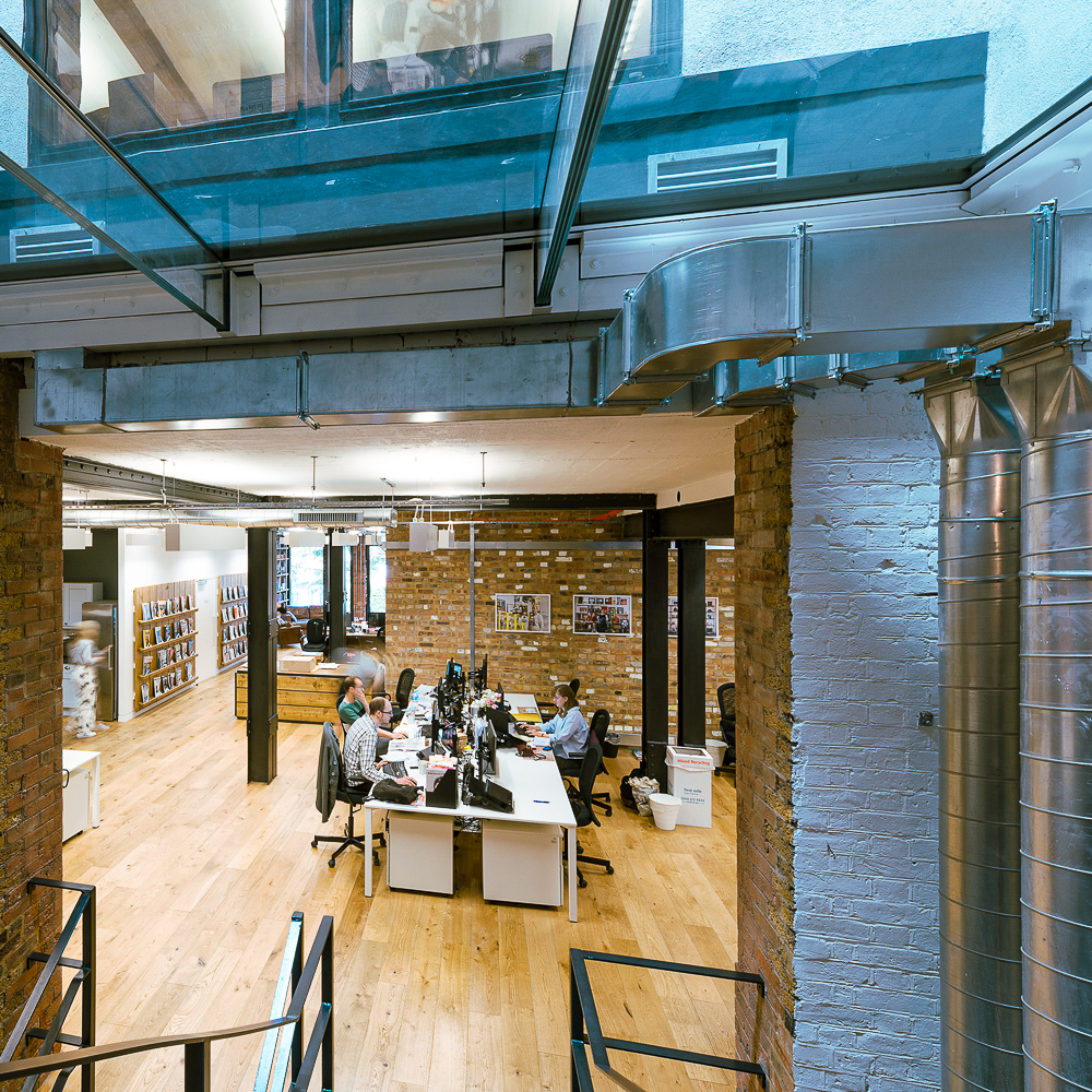 Open-plan office with people working at desks, featuring exposed brick walls, large windows, wooden floors, and ductwork. Shelves with books line the back wall.