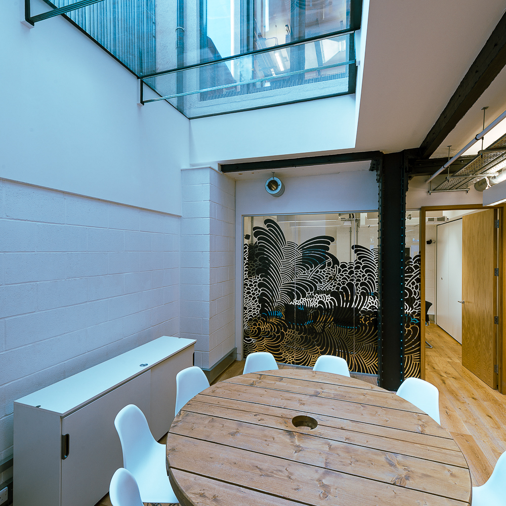 A modern office space with a wooden round table surrounded by white chairs. Glass partitions with abstract patterns separate the area. Skylight and wood accents provide natural light and warmth.