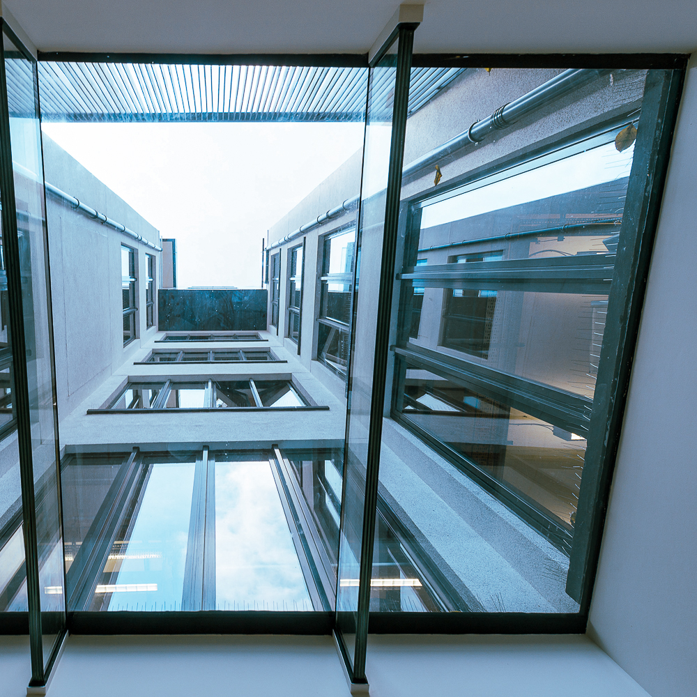 Upward view of a narrow building courtyard with reflective glass windows, framed by a clear glass ceiling, showing a cloudy sky.