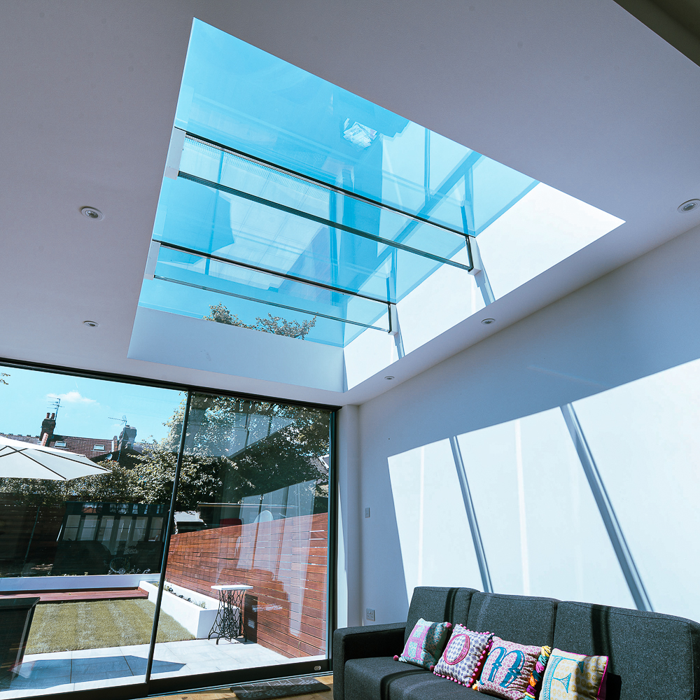 A modern living room with a large glass skylight and sliding glass doors, allowing sunlight to illuminate the space. A gray couch with colorful pillows is against the wall.