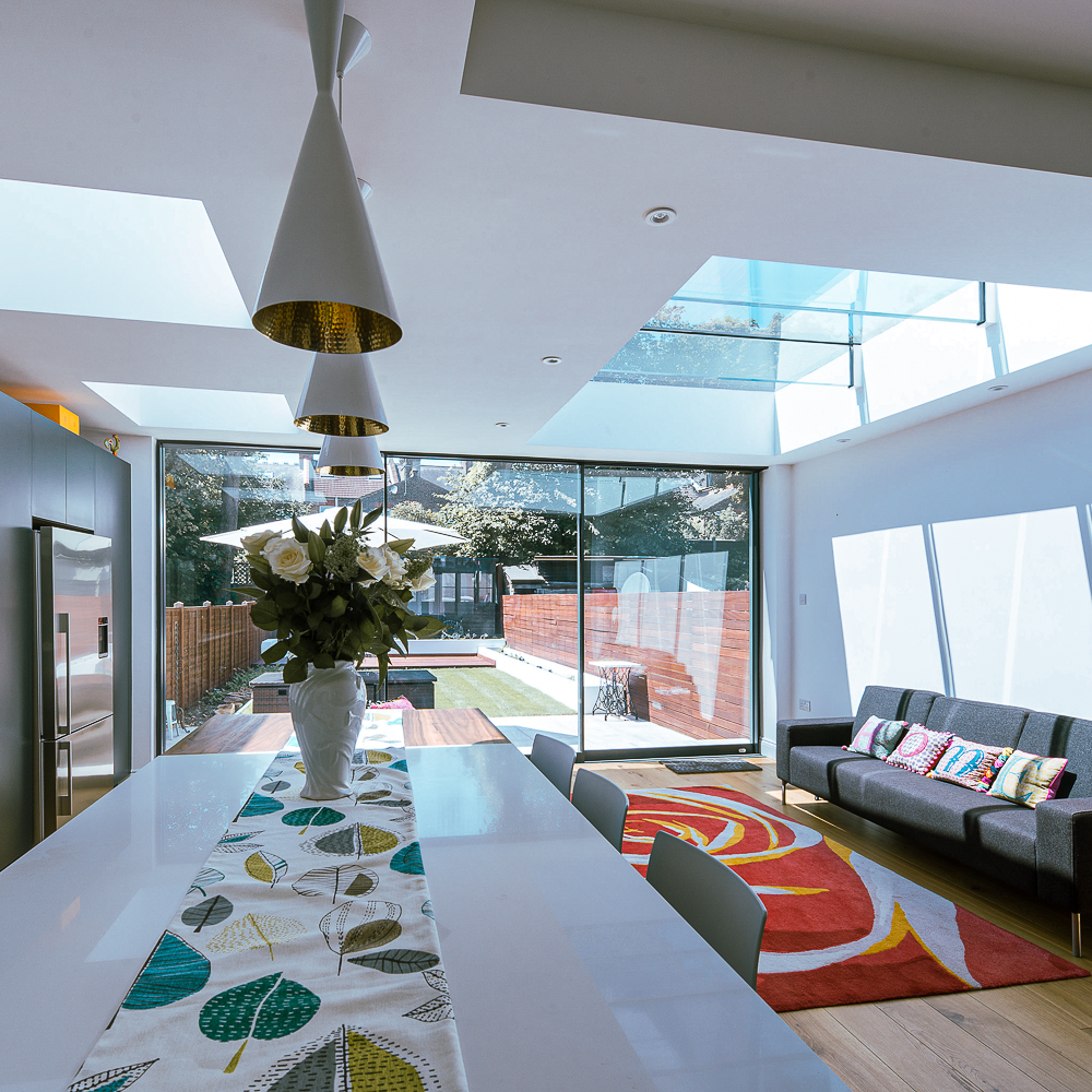 Modern kitchen and living area with a long table, floral centerpiece, colorful rug, sofa, and large glass doors opening to a garden.