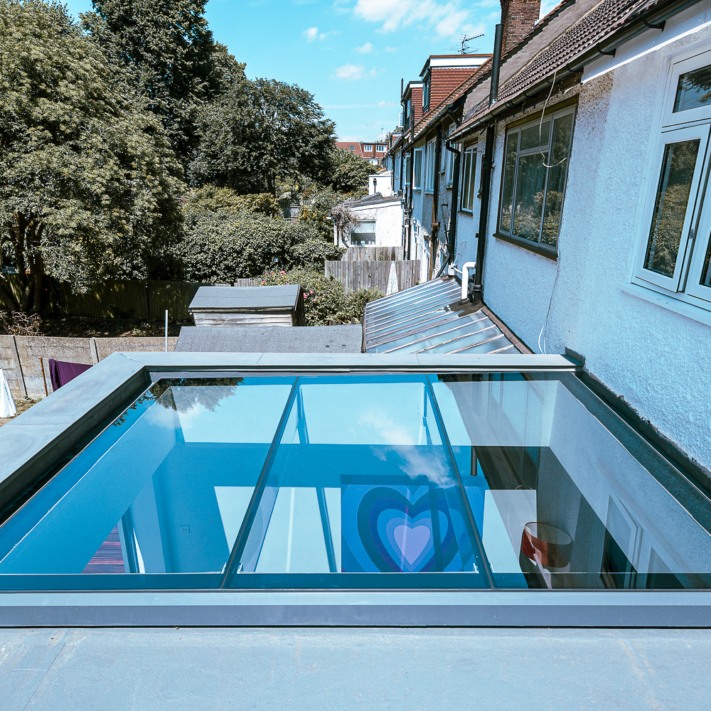 A rooftop with a large glass skylight reflects the sky. Behind are trees and the upper floors of adjacent houses. A heart-shaped decoration is visible indoors through the skylight.