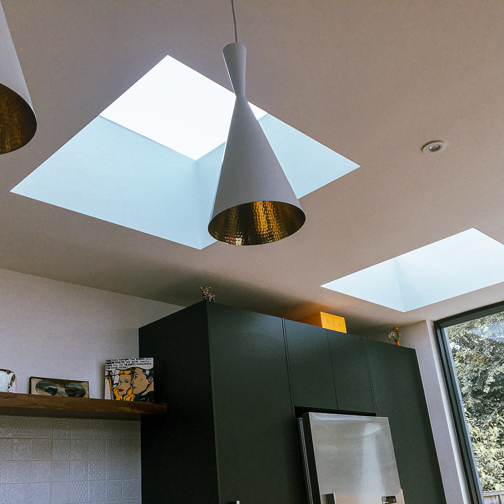 Modern kitchen with two skylights, geometric pendant light, dark cabinetry, refrigerator, and a shelf holding books and framed art. Large window offers a view of trees outside.
