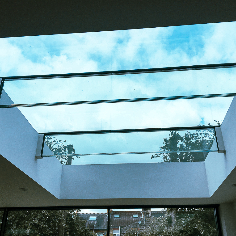 View of a large glass skylight with a blue sky and clouds above, framed by white walls and overlooking trees and rooftops.