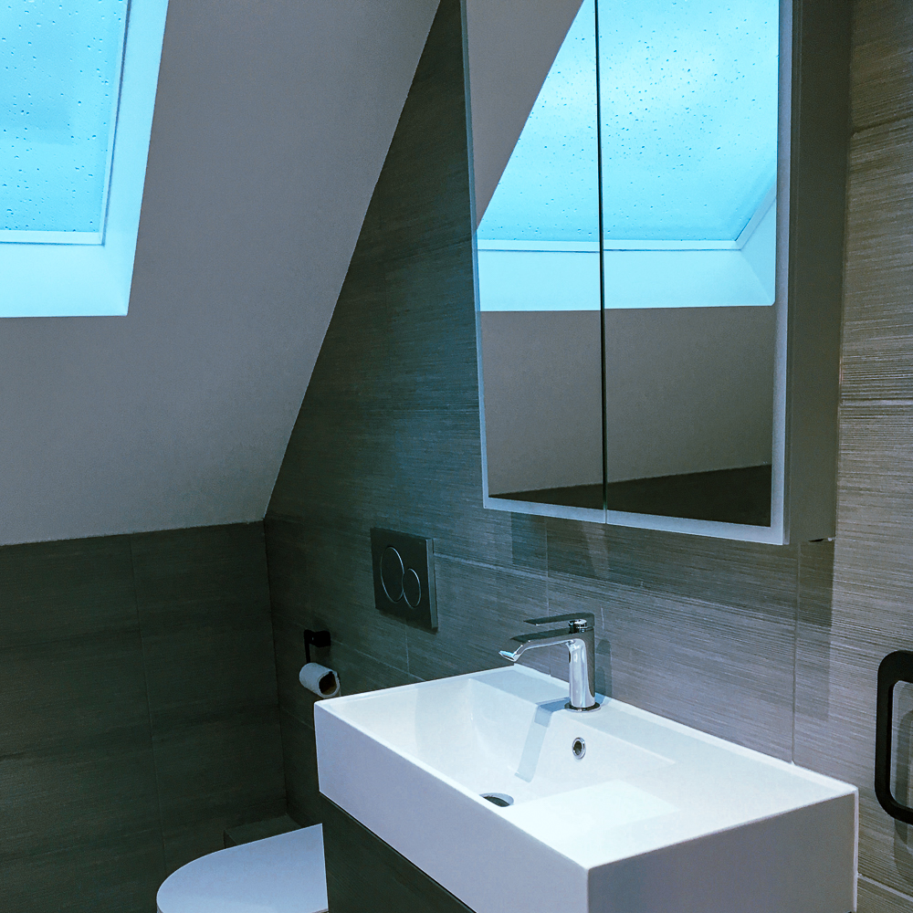 Bathroom with a small white sink, a mirror cabinet, and a toilet. Slanted ceiling with a skylight casting blue light on the gray tiled walls.