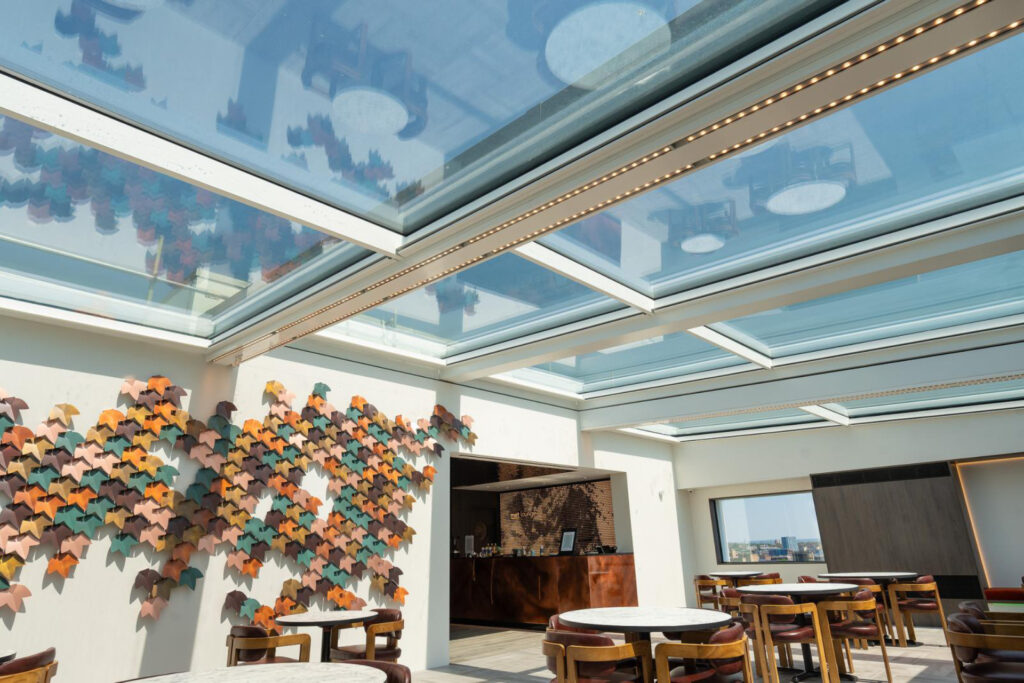 A modern indoor seating area with round tables and chairs. A wall is decorated with colorful, abstract art shaped like birds. The space features large, glass skylights letting in natural light.