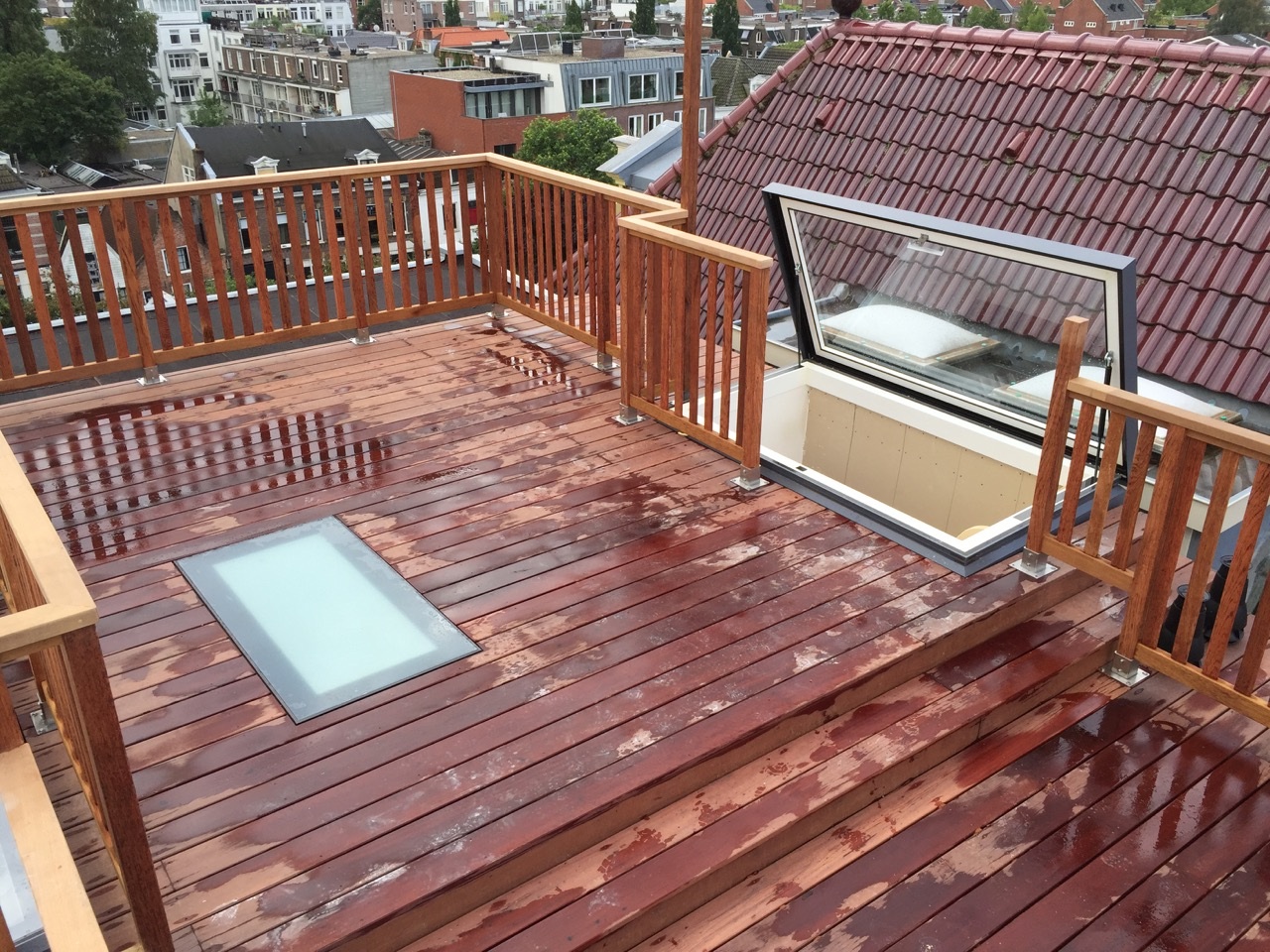 Rooftop terrace with wet, wooden decking and railings, surrounded by buildings. A skylight and a hatch are visible on the terrace.