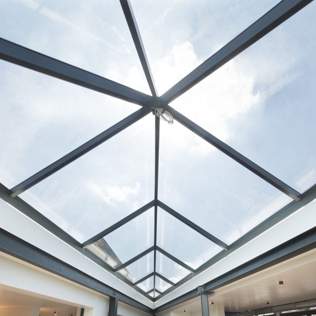 View from below of a modern glass ceiling with a metal frame, showcasing a clear blue sky and clouds.