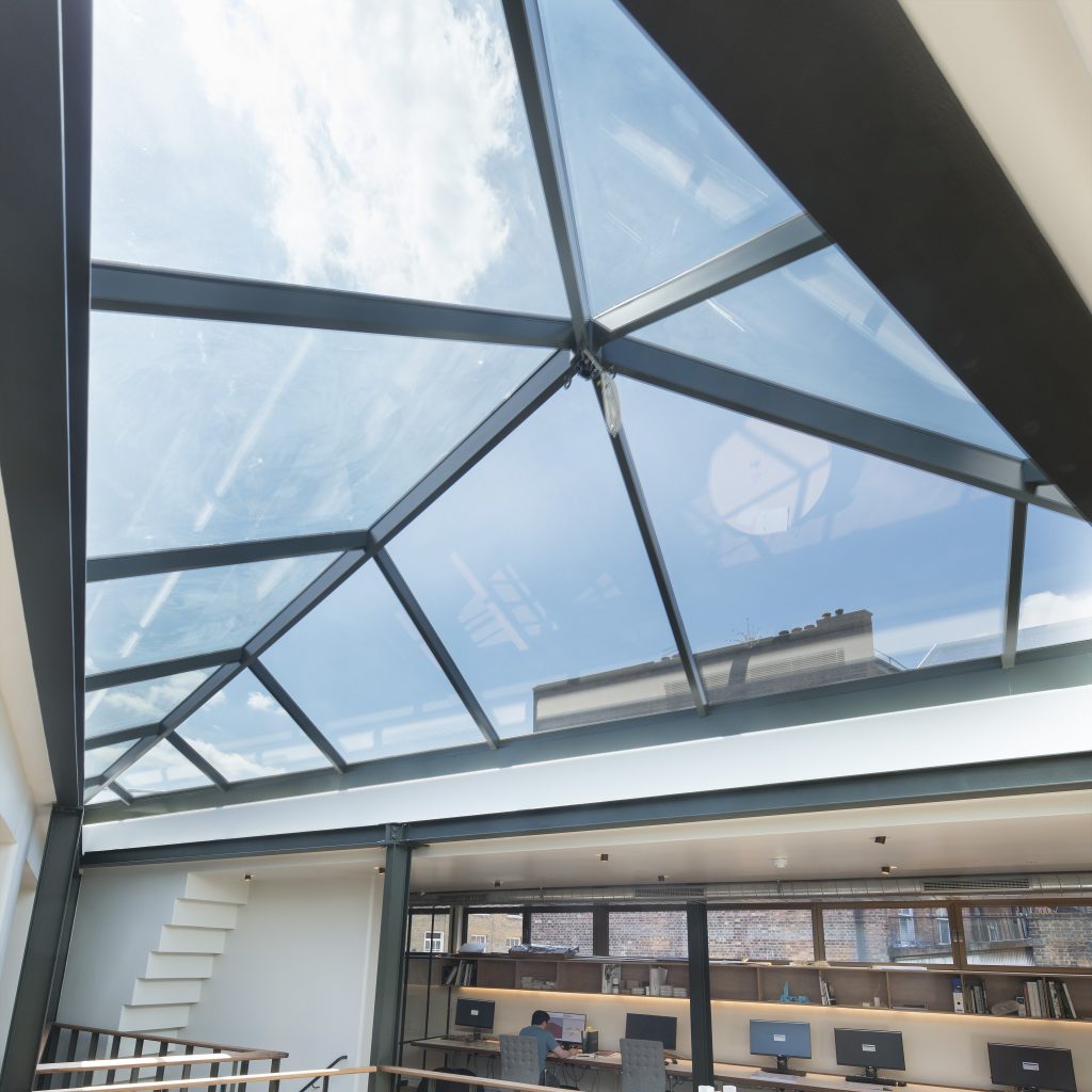 Interior space with a glass ceiling showing a view of the sky. Below, there are desks and chairs against a brick wall.