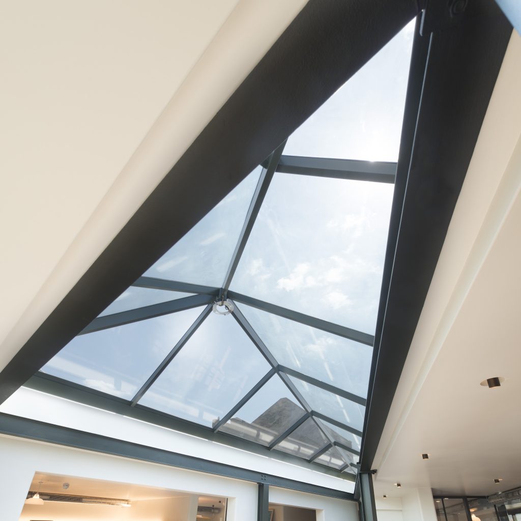 A triangular glass skylight with black metal frames is set in a white ceiling, allowing sunlight and blue sky to be visible.