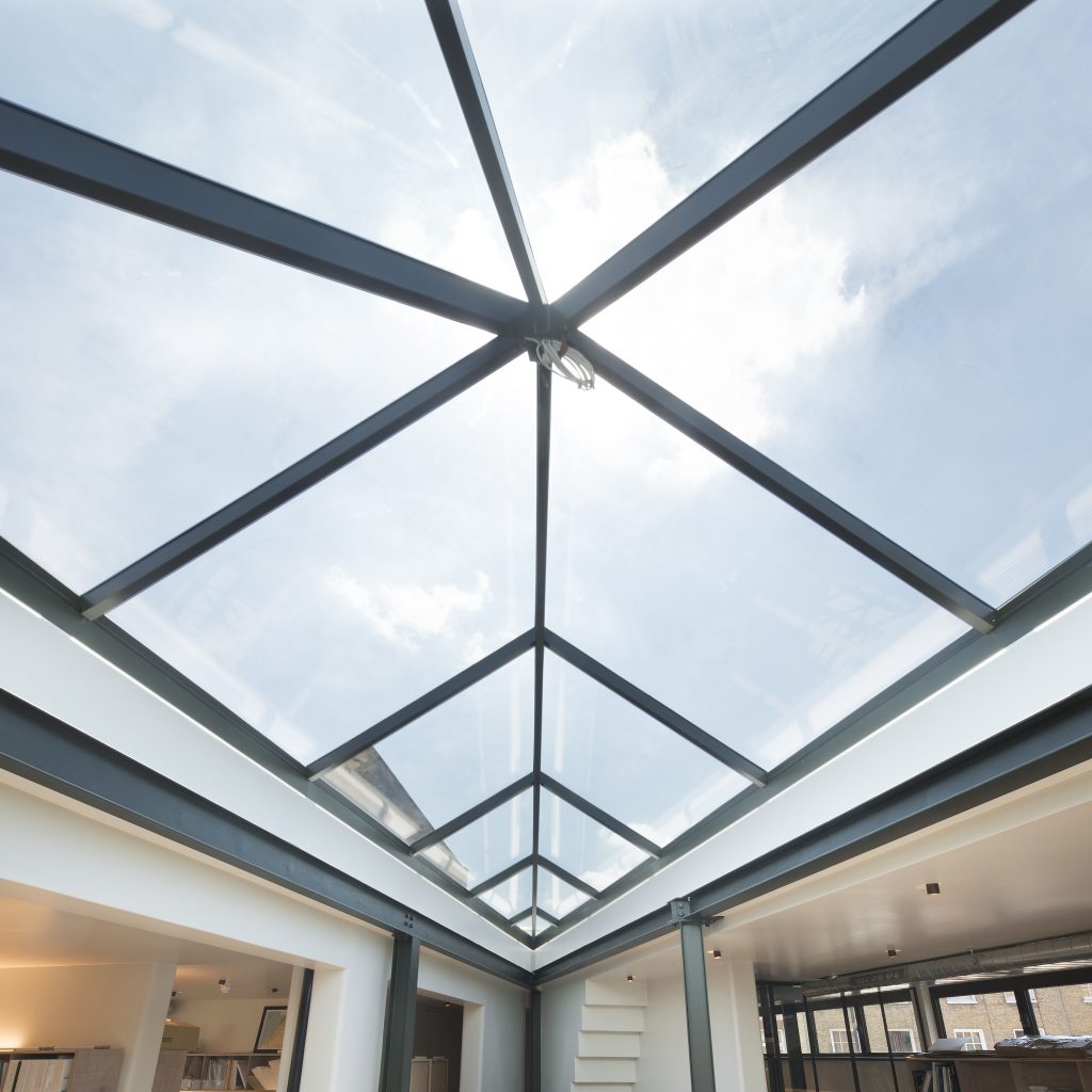 A modern office space with a large glass ceiling, allowing natural light to illuminate the room. Desks and shelves are visible below.