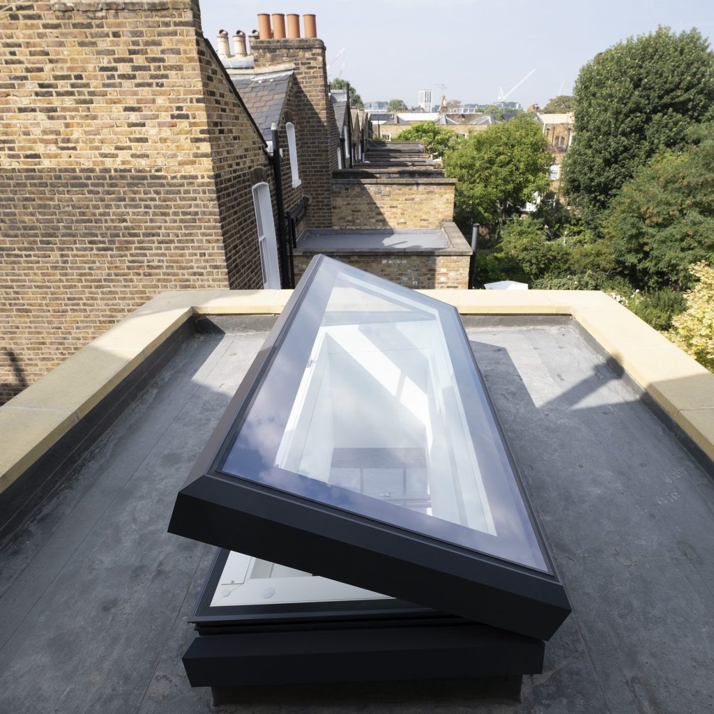 A rooftop with an open skylight, surrounded by brick buildings and overlooking trees in the background.