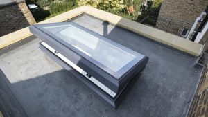 A hinged skylight installed on a flat rooftop with surrounding brick walls and trees in the background.