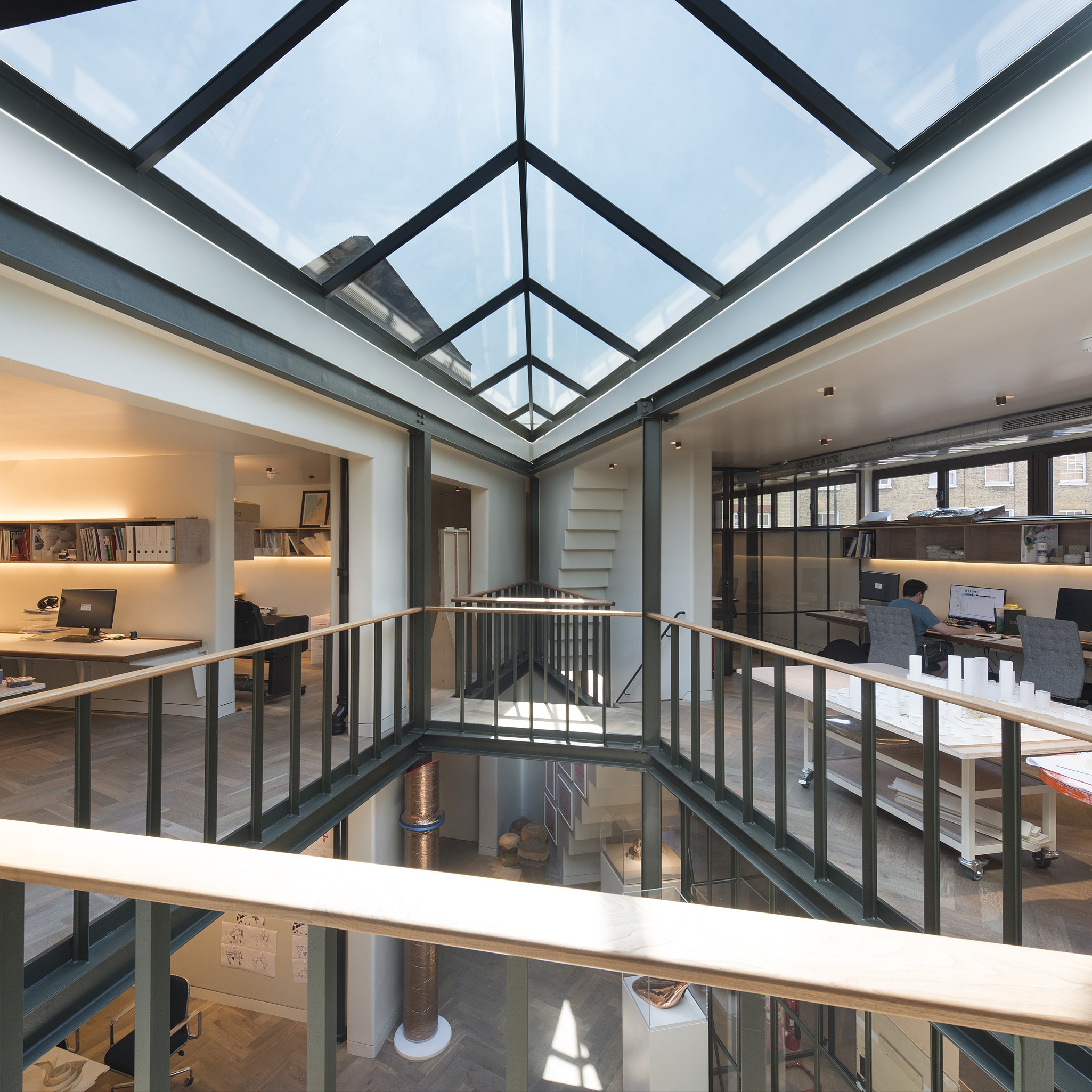 Modern office interior with glass ceiling and open layout, featuring desks, shelves with books, and a central stairway. Natural light enters through the ceiling, illuminating the workspace.