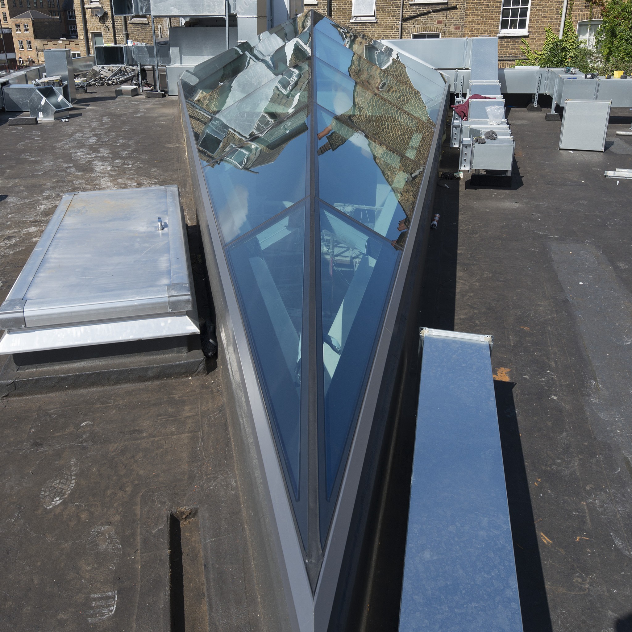 A rooftop with metal structures and a large, triangular reflective glass installation surrounded by brick buildings.