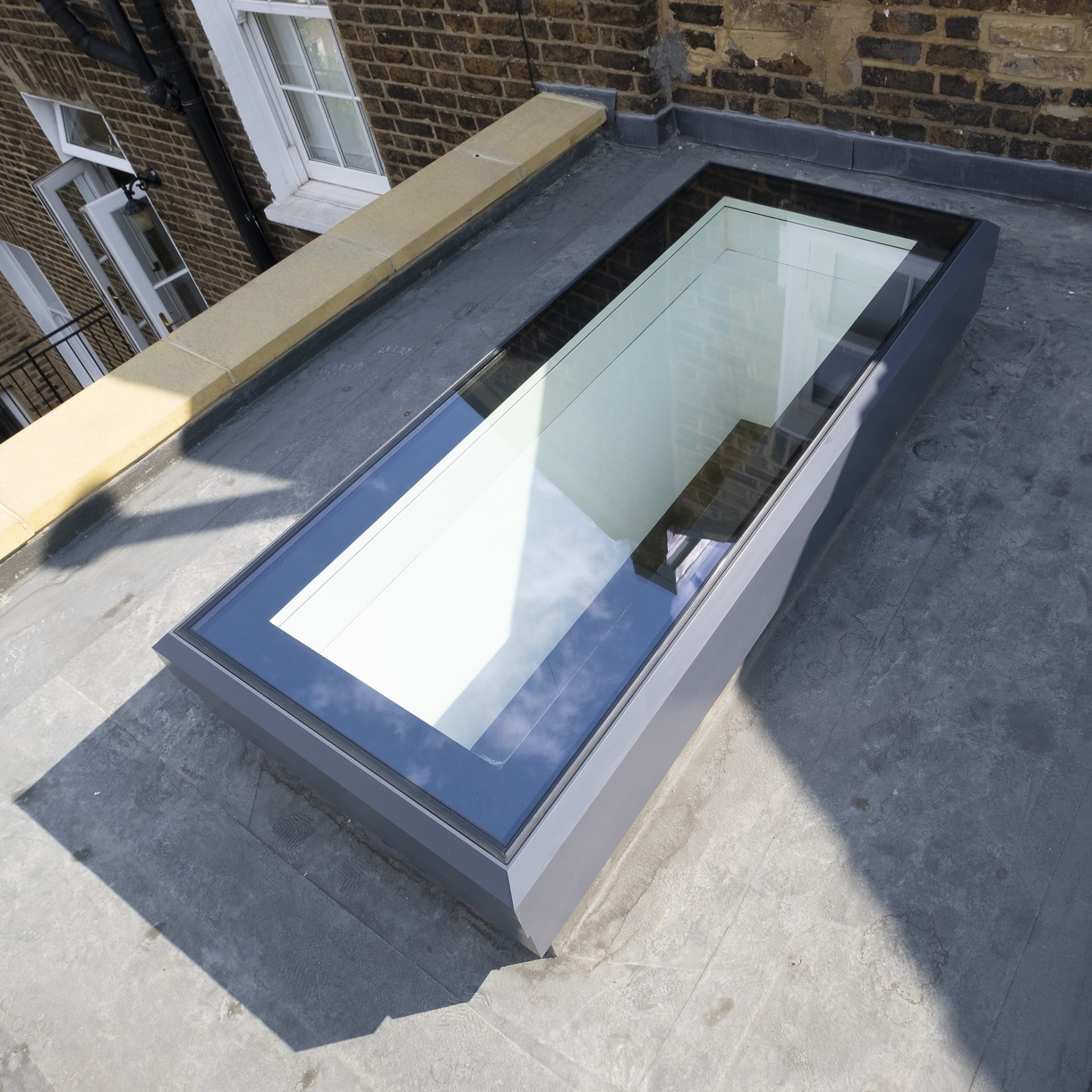 Rectangular skylight with a blue frame installed on a flat rooftop, reflecting the sky and surrounding buildings.