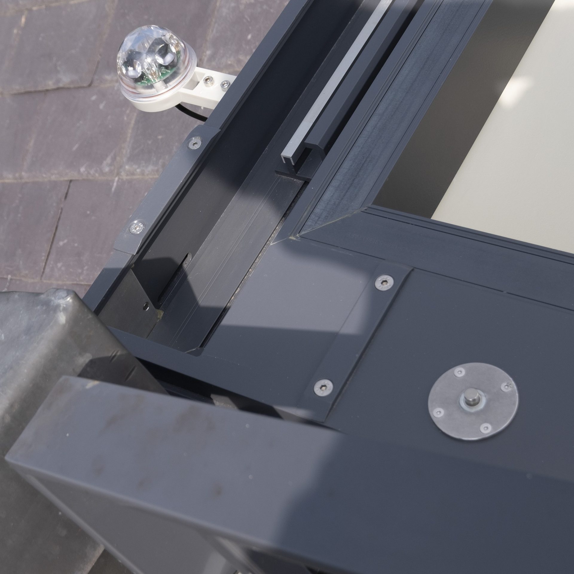 Close-up of a modern window opening mechanism with metallic and glass components, showing screws and brackets, against a tiled roof background.