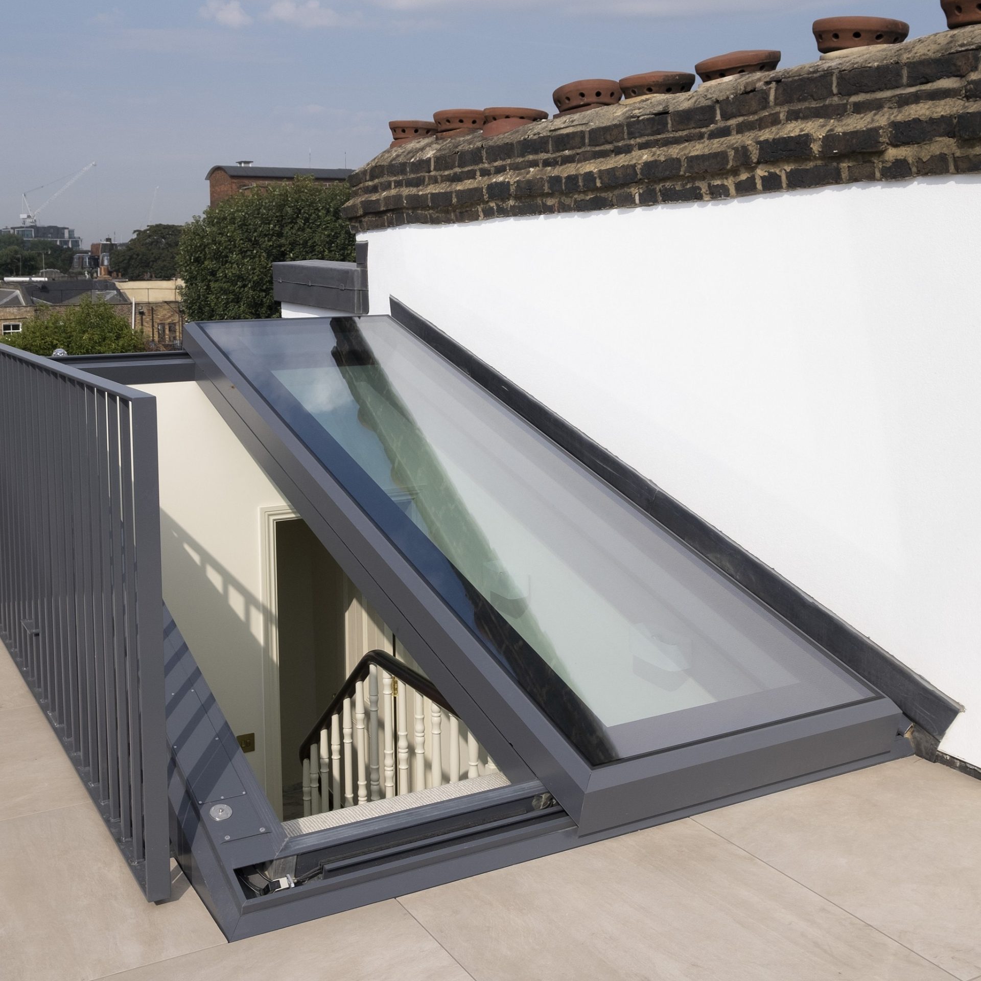 A rooftop glass skylight hatch is open, revealing a staircase leading down. The sky is clear, and rooftops are visible in the background.