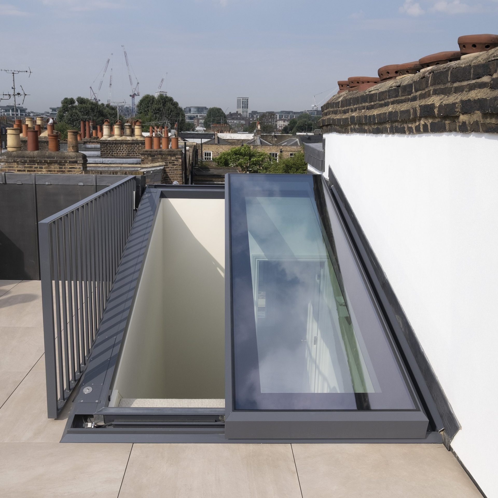 A rooftop with a sliding glass skylight partially open, revealing stairs leading down. Brick chimney stacks and distant cityscape are visible under a clear blue sky.