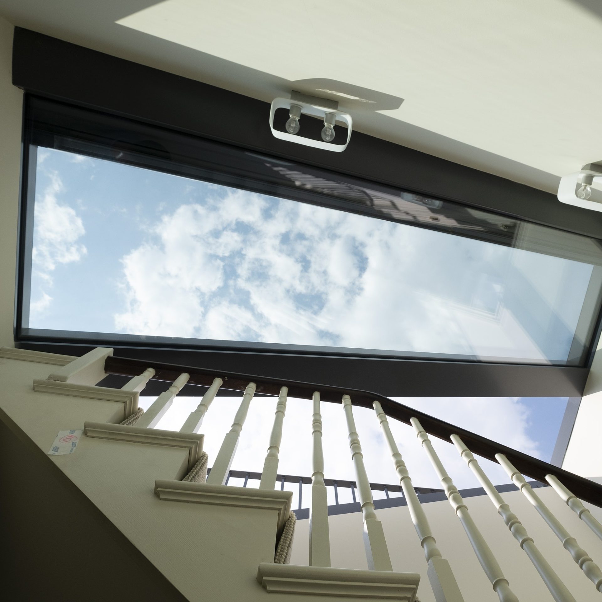 View from below a modern staircase with white railings, looking up through a window displaying a clear blue sky with scattered clouds.