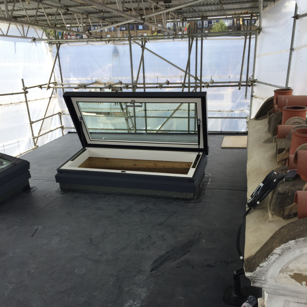 An open skylight on a roof surrounded by scaffolding and protective covering, with stacks of roof tiles nearby.
