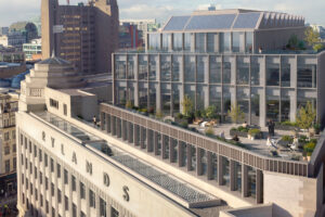 A modern glass building with rooftop terraces sits atop the historic Rylands building in a cityscape. People are seen on the terraces among trees and seating areas.