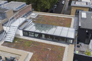 A rooftop with a glass structure and a green roof featuring sparse vegetation, surrounded by gravel and rooftop equipment, with a metal staircase on the left.