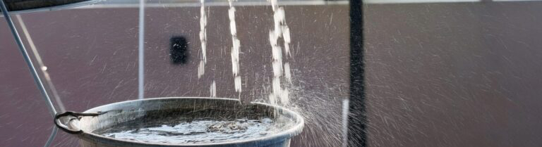Water is being poured into a metal bucket with several holes, causing water to leak out in streams.
