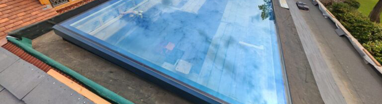 Rectangular glass rooflight installed on a flat roof, reflecting the sky and surroundings, with roofing materials and greenery visible nearby.
