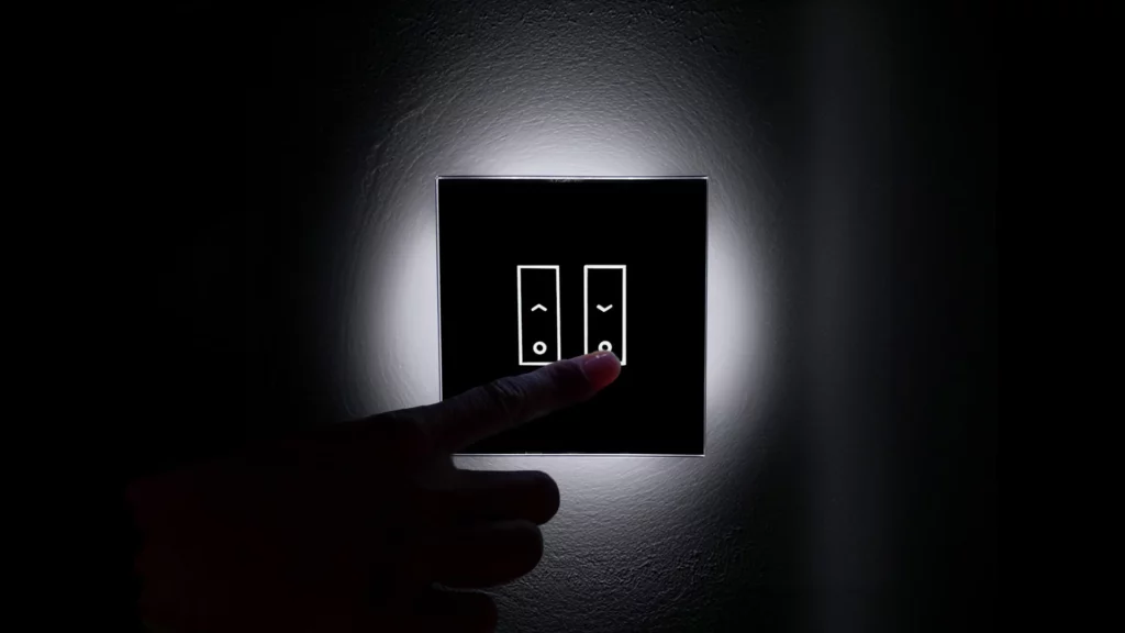 A hand is about to press a button on a backlit, modern black light switch panel mounted on a wall.