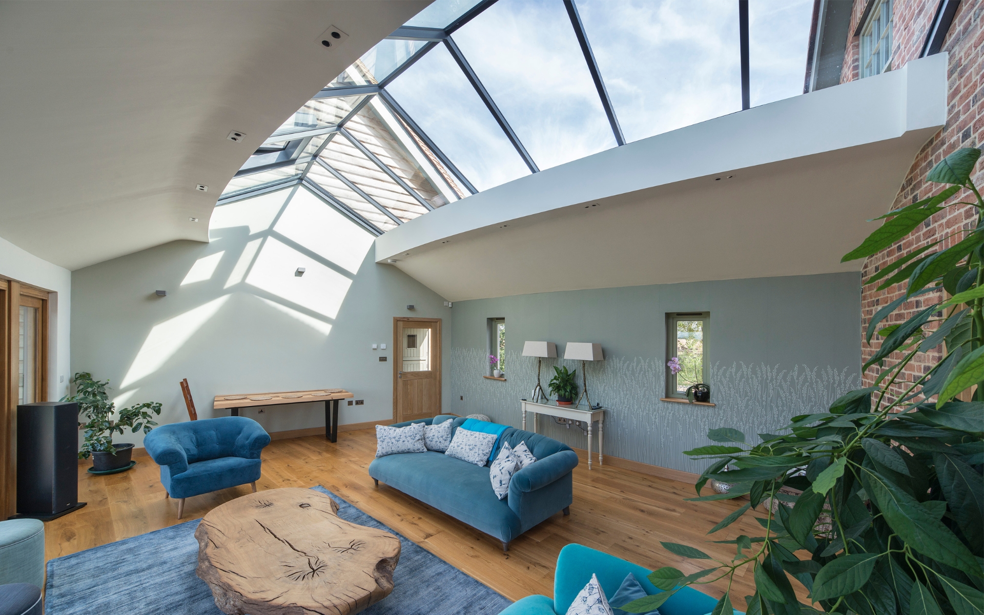 A modern living room with a glass ceiling, blue sofas, a wooden coffee table, houseplants, and large windows letting in natural light.