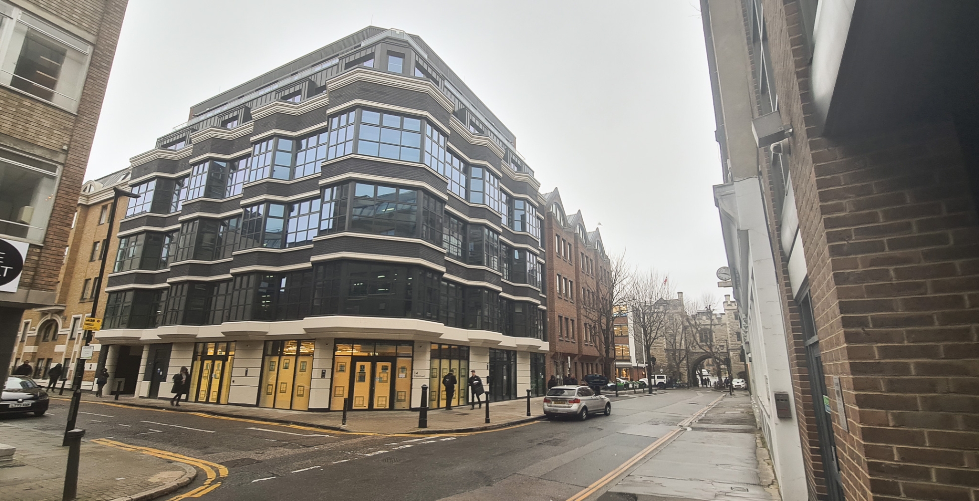 Modern multi-storey building with large glass windows and a striking three-wall box rooflight at a street corner on an overcast day; few cars and pedestrians are visible.