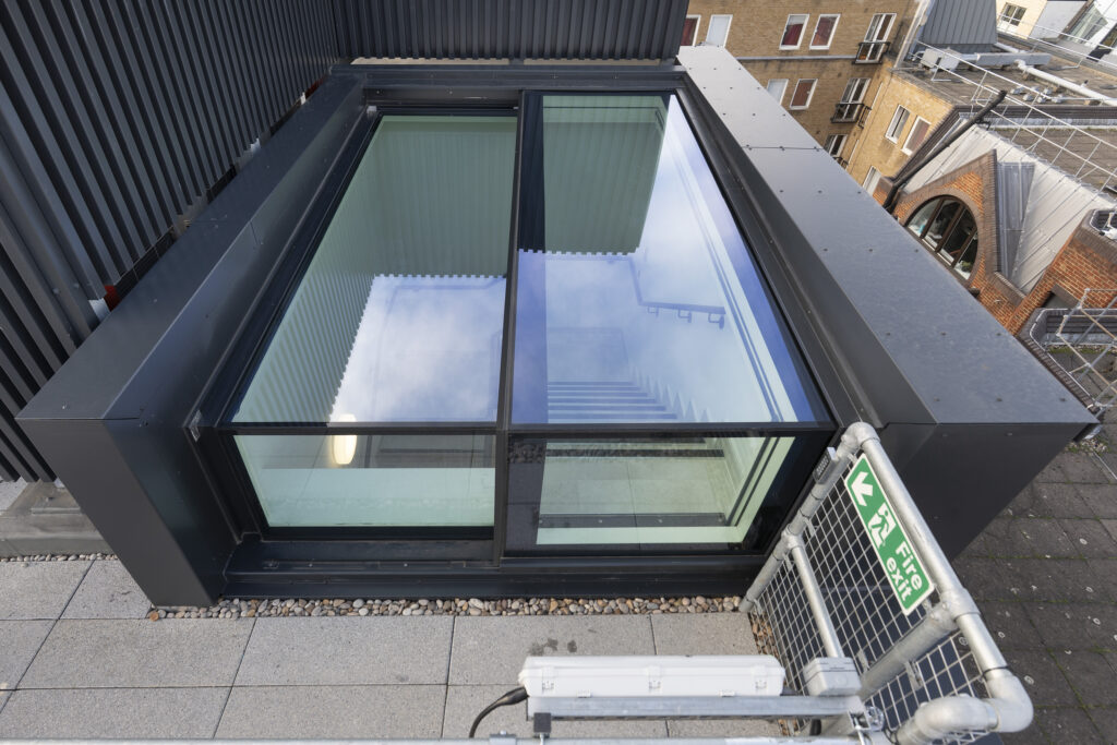 A rooftop features a large three wall box rooflight revealing a stairwell below, surrounded by metal railings and adjacent buildings.