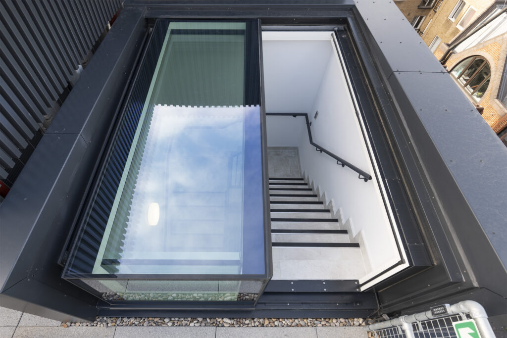 A modern three-wall box rooflight next to a staircase leading down into a white-walled basement with a black handrail.