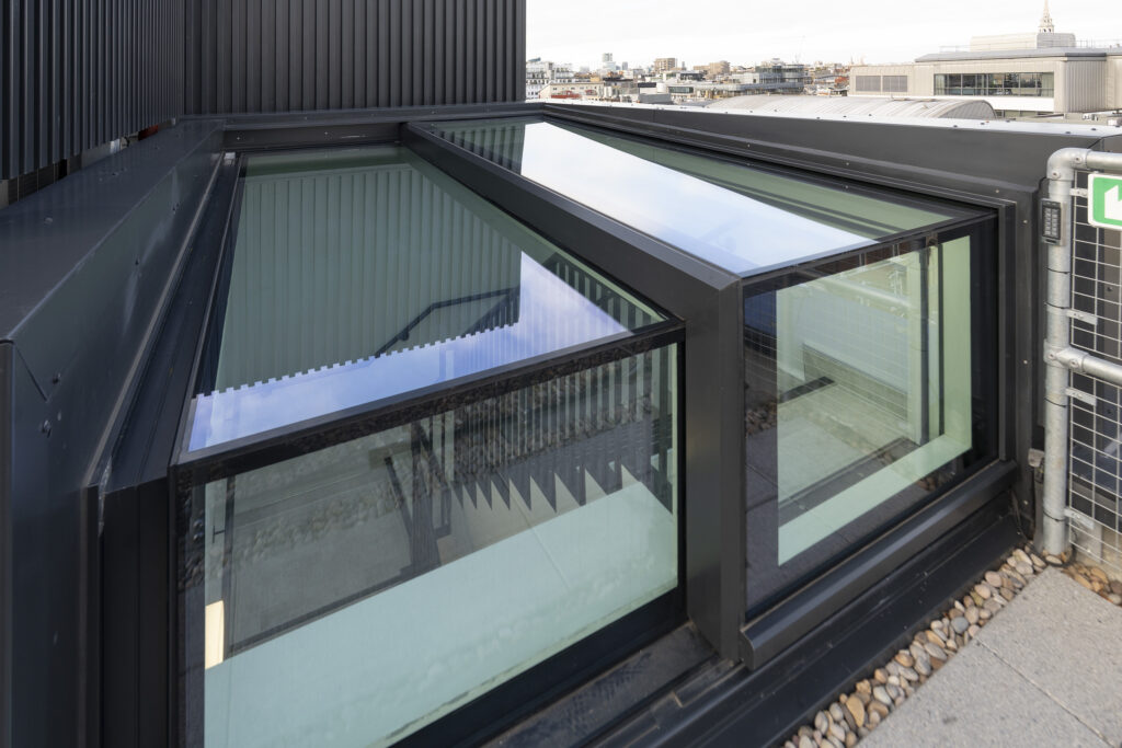 A modern three-wall box rooflight with large glass panes, set at an angle, bordered by a black metal frame, next to a gravel-lined walkway.