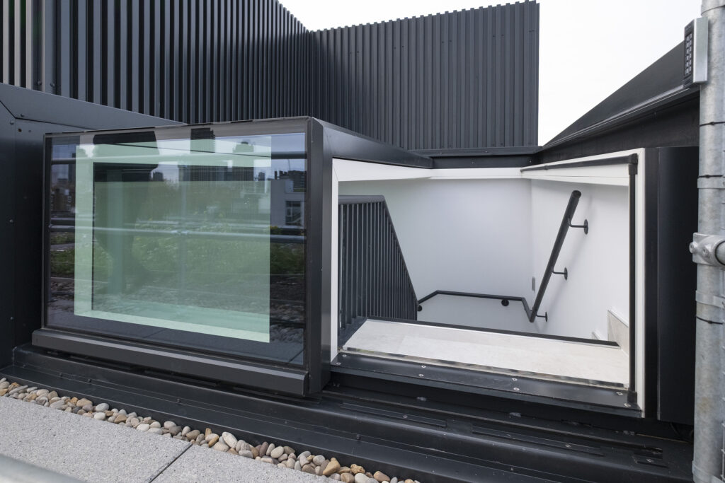 Glass-enclosed rooftop stairwell entrance featuring a three-wall box rooflight, with black metal framing and white interior walls, leading down a staircase with sleek black handrails.