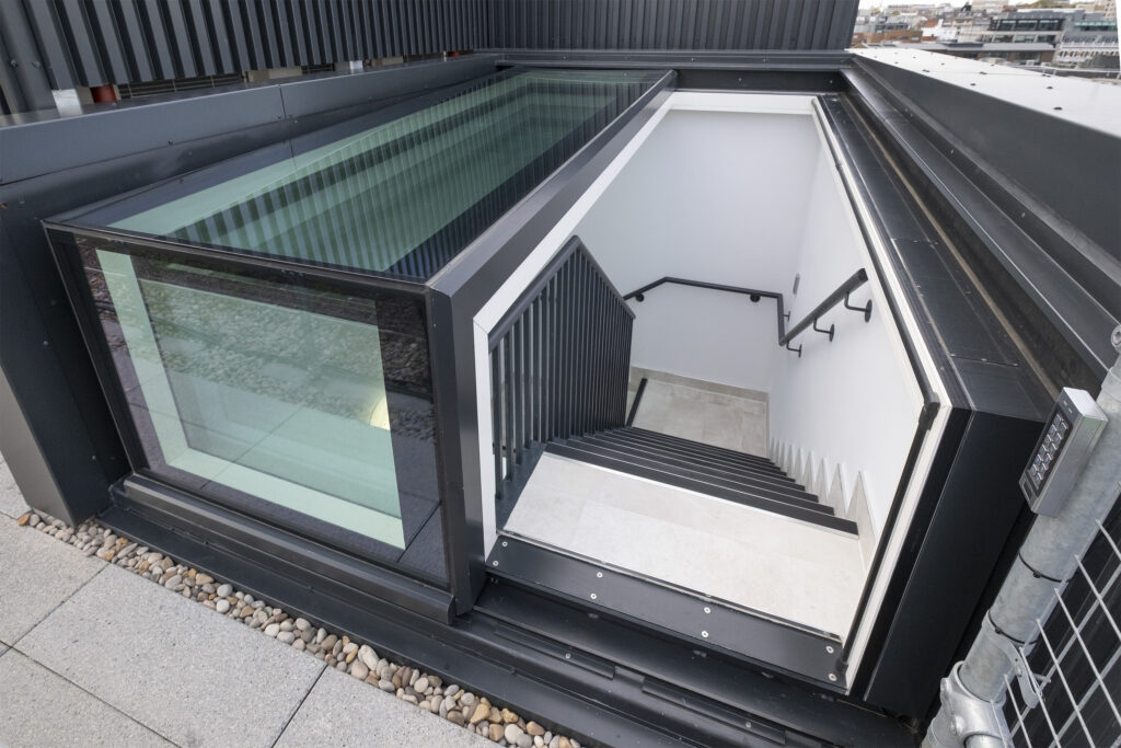 A modern staircase leading down, featuring black handrails, white walls, and a striking three wall box rooflight that floods the space with natural light.