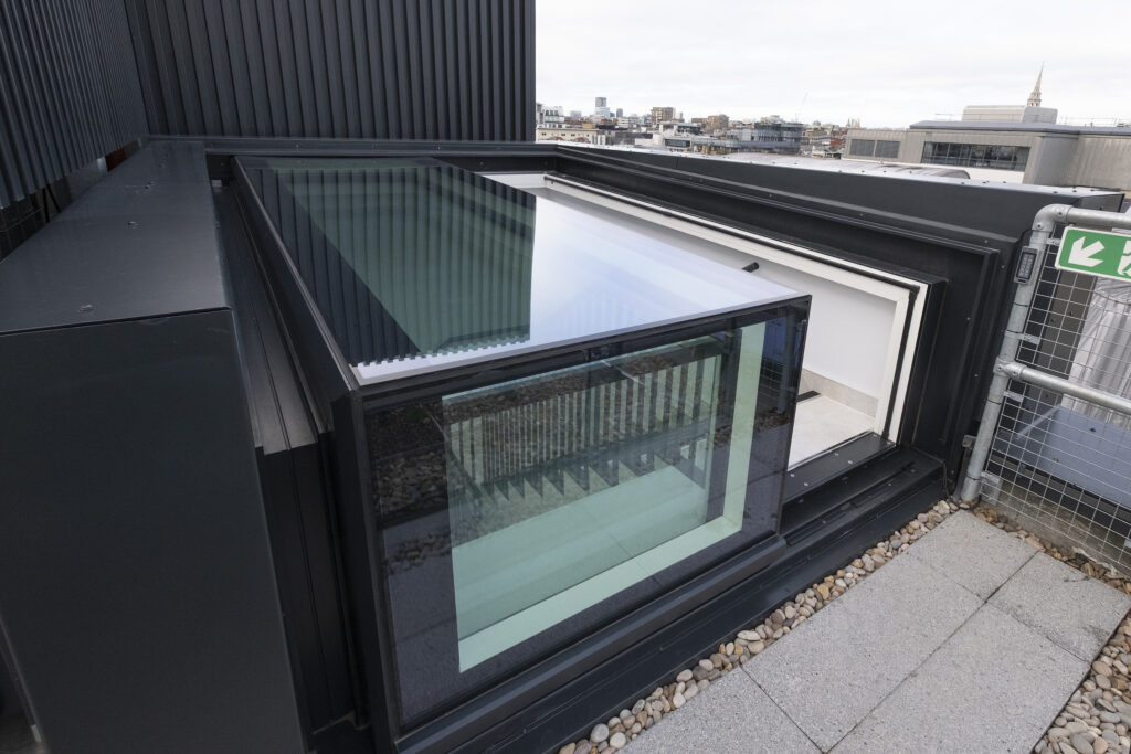 A modern rooftop glass sliding door partially open, leading from an indoor space with a three-wall box rooflight to an outdoor terrace with gravel and paving stones, overlooking a cityscape.