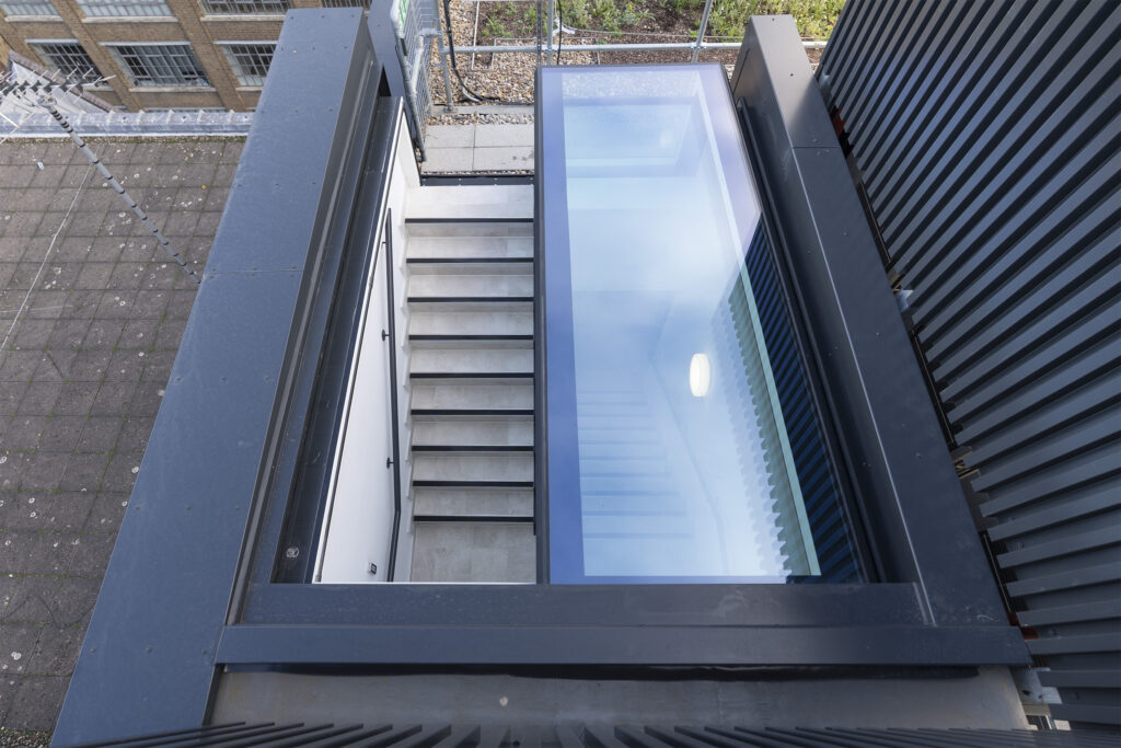 View from above of a three wall box rooflight with a staircase surrounded by modern buildings, featuring a nearby that brightens the contemporary space.
