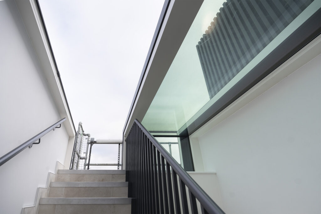 Stairway with beige steps, black railings and white walls under a striking three wall box rooflight.