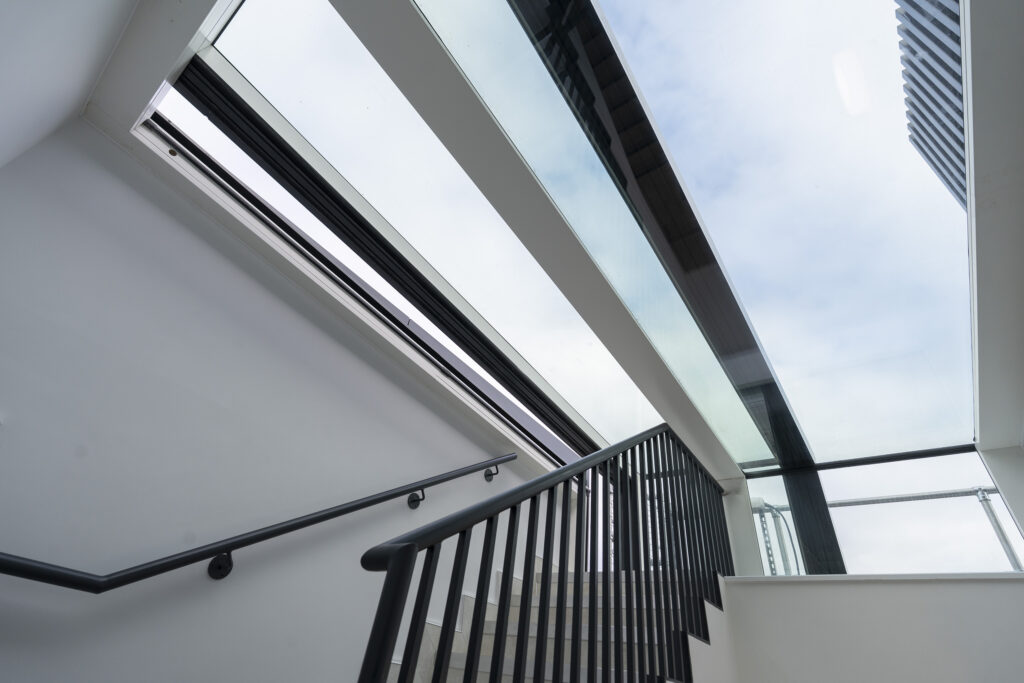 A modern indoor staircase with black railings leads up to a landing, featuring large windows and a three-wall box rooflight that offers a view of a tall building outside.