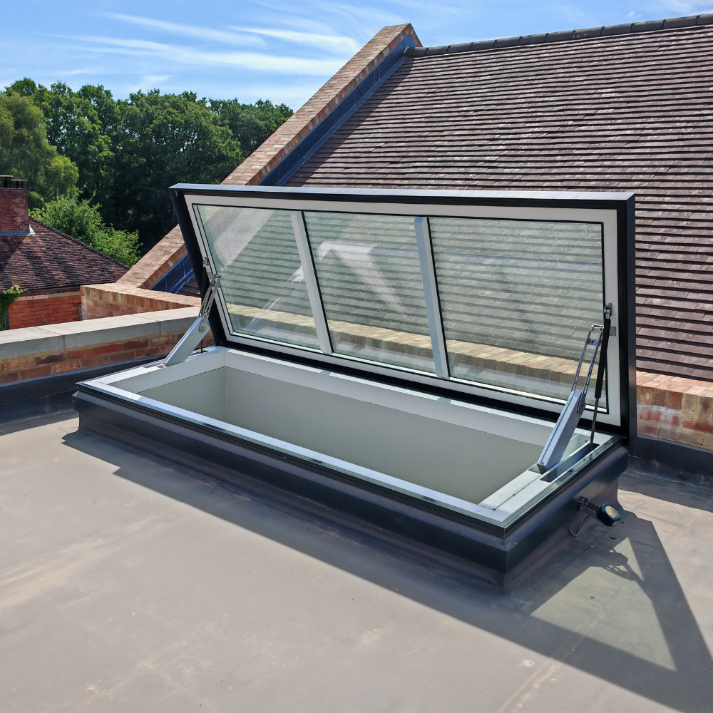 Large Access Hatch with Weather Sensor A large access hatch with a white frame and metal hinges stands open on a flat rooftop, revealing the rectangular glass roof as pitched roof buildings and trees appear in the background.