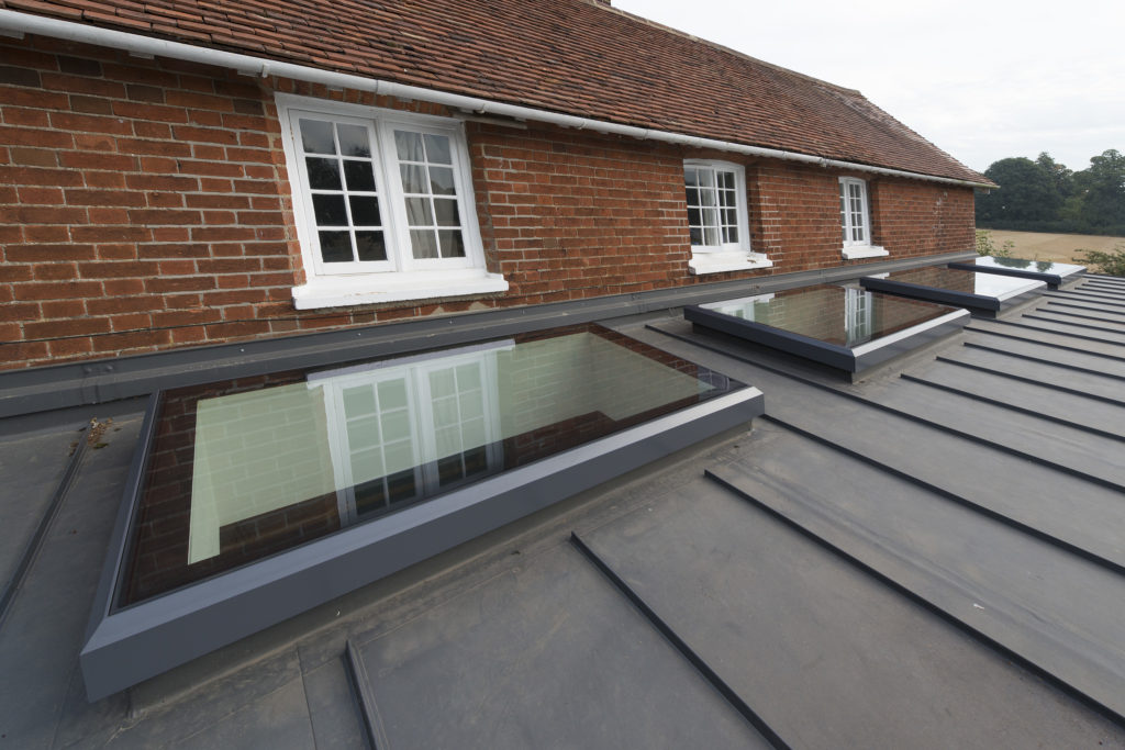 Flat glass rooflights installed on a metal roof beneath brick walls with white-framed windows, showcasing the difference between framed vs frameless rooflights. Trees and fields are visible in the background.